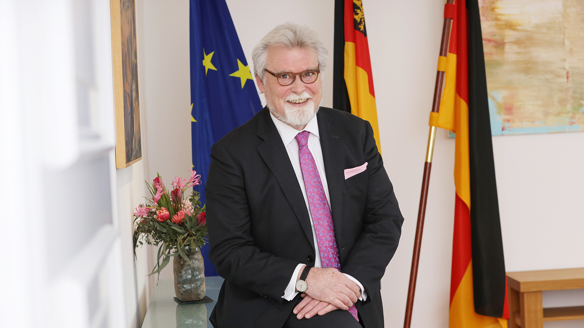 Justizminister Herbert Mertin auf einem Sideboard sitzend, im Hintergrund Fahnen (v.l.n.r.: Europa, Rheinland-Pfalz, Deutschland)