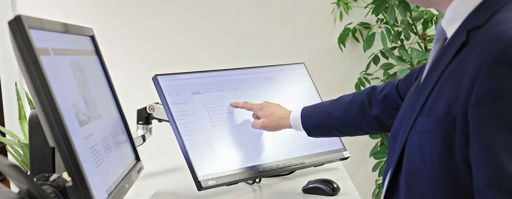 Eine männliche Person stehend vor einem Schreibtisch mit zwei Monitoren, einem Telefon und einer Tastatur. Männliche Person bedient einen Touch-Bildschirm