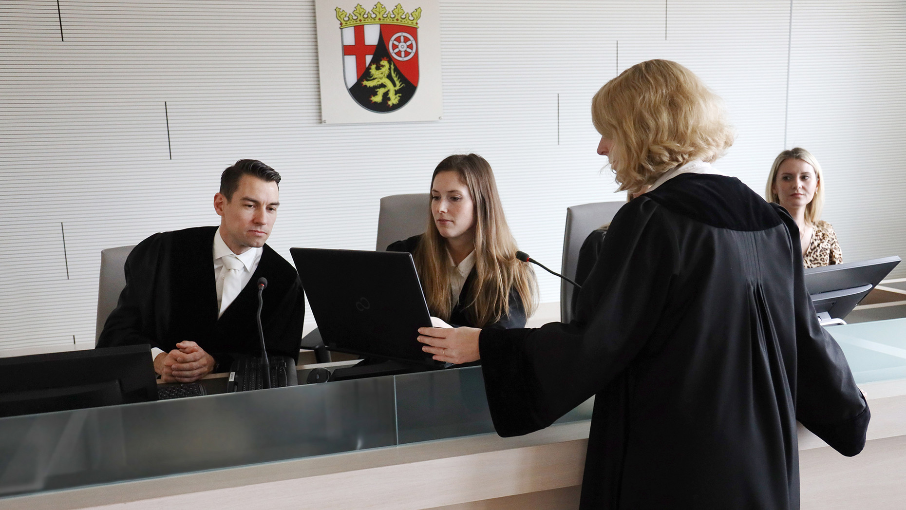 Staatsanwältin zeigt einem Richter und einer Richterin an der Richterbank etwas auf ihrem Laptop