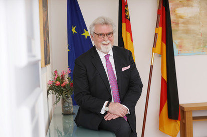 Justizminister Herbert Mertin auf einem Sideboard sitzend, im Hintergrund Fahnen (v.l.n.r.: Europa, Rheinland-Pfalz, Deutschland)