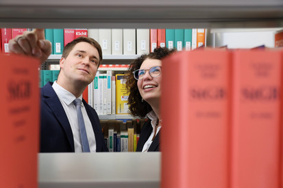 Eine männliche und eine weibliche Person, welche durch eine Bücherlücke in der Bibliothek zu sehen sind und in ein oberes Regal schauen.