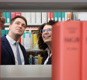 Eine männliche und eine weibliche Person, welche durch eine Bücherlücke in der Bibliothek zu sehen sind und in ein oberes Regal schauen.